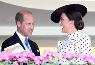 Prince William wearing a suit looking at Kate Middleton who is laughing and wearing a white polkadot dress and large hat