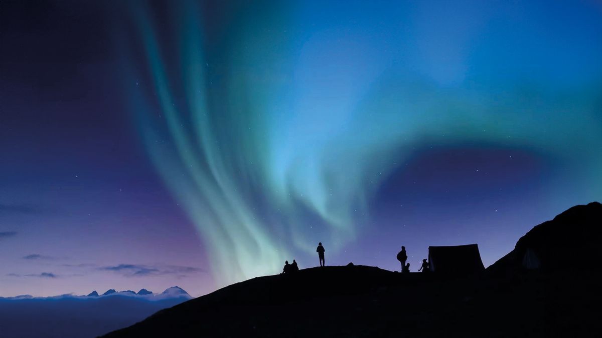 At night, a silhouette of a mountain and people are visible against a starry blue sky. The sky glows with purple and turquoise hues due to the northern lights