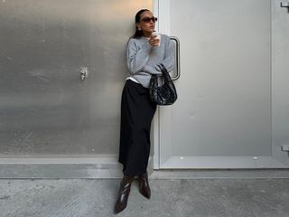 Fashion influencer @ingridedvinsen wearing an elegant everyday outfit with a black skirt, knee-high boots, a woven leather bag, gray sweater worn over a white t-shirt, and sunglasses.