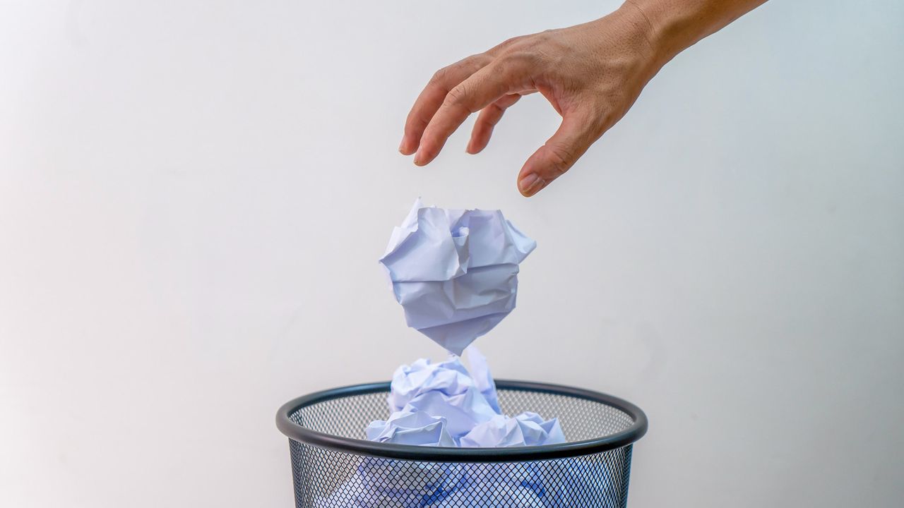 A hand throws a balled up piece of paper in a waste basket.