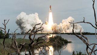 a space shuttle launches in the distance with large plumes of smoke on either side. The foreground shows flora near a body of water, which reflects the rocket engine flames.