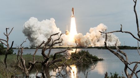 a space shuttle launches in the distance with large plumes of smoke on either side. The foreground shows flora near a body of water, which reflects the rocket engine flames.