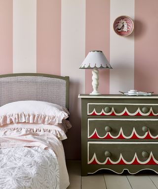 Colorful and contemporary bedroom with pink striped walls, bed with upholstered headboards, painted patterned chest of draws, decorated with table lamp and ornaments