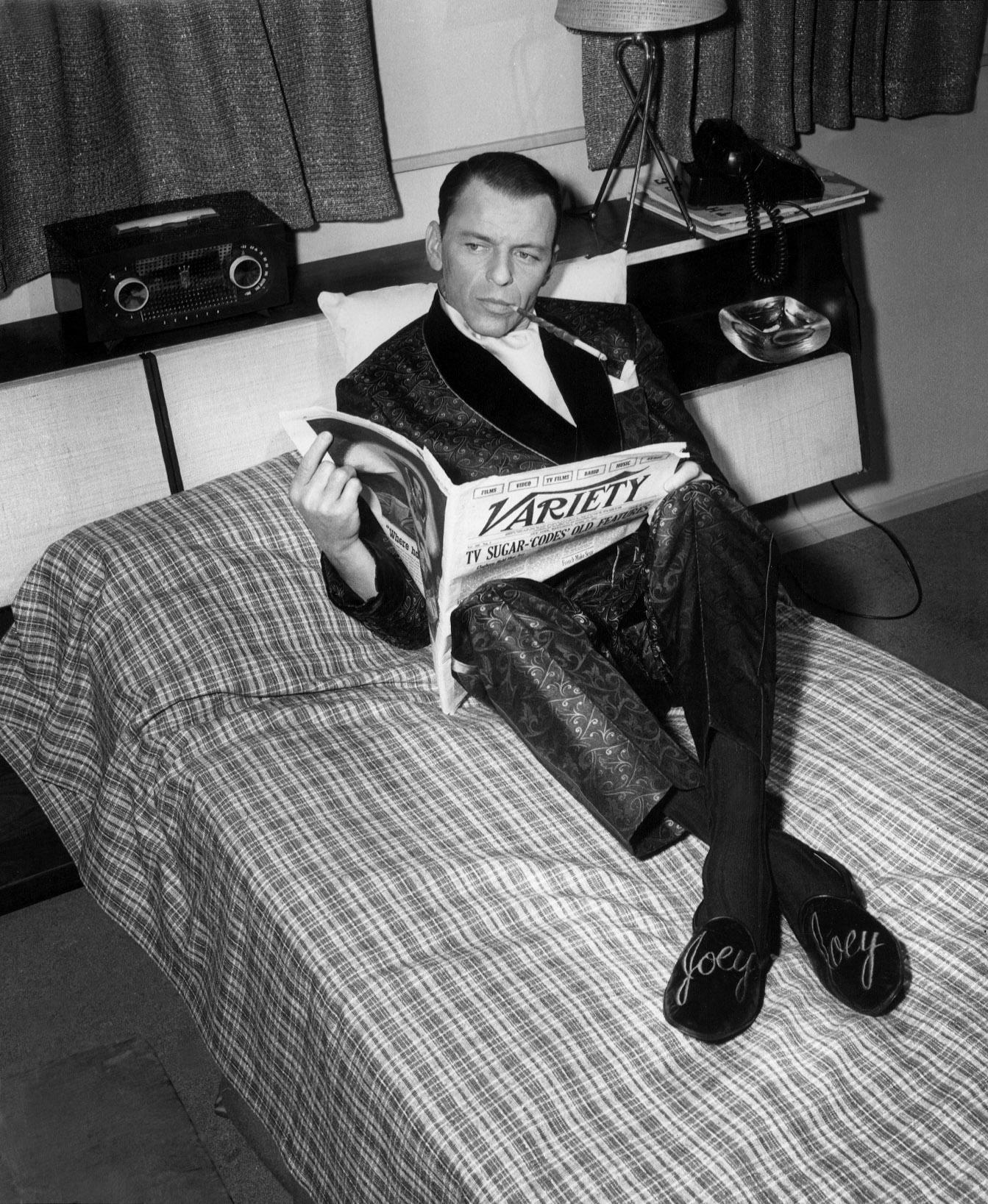 Frank sinatra sat on a bed reading variety while wearing a smoking jacket and slippers