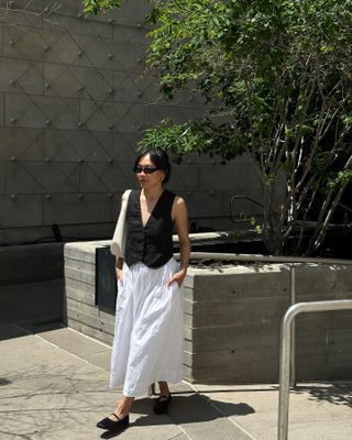 a photo of a woman wearing sunglasses, a white hobo bag, a black vest, a white skirt, and ballet flats