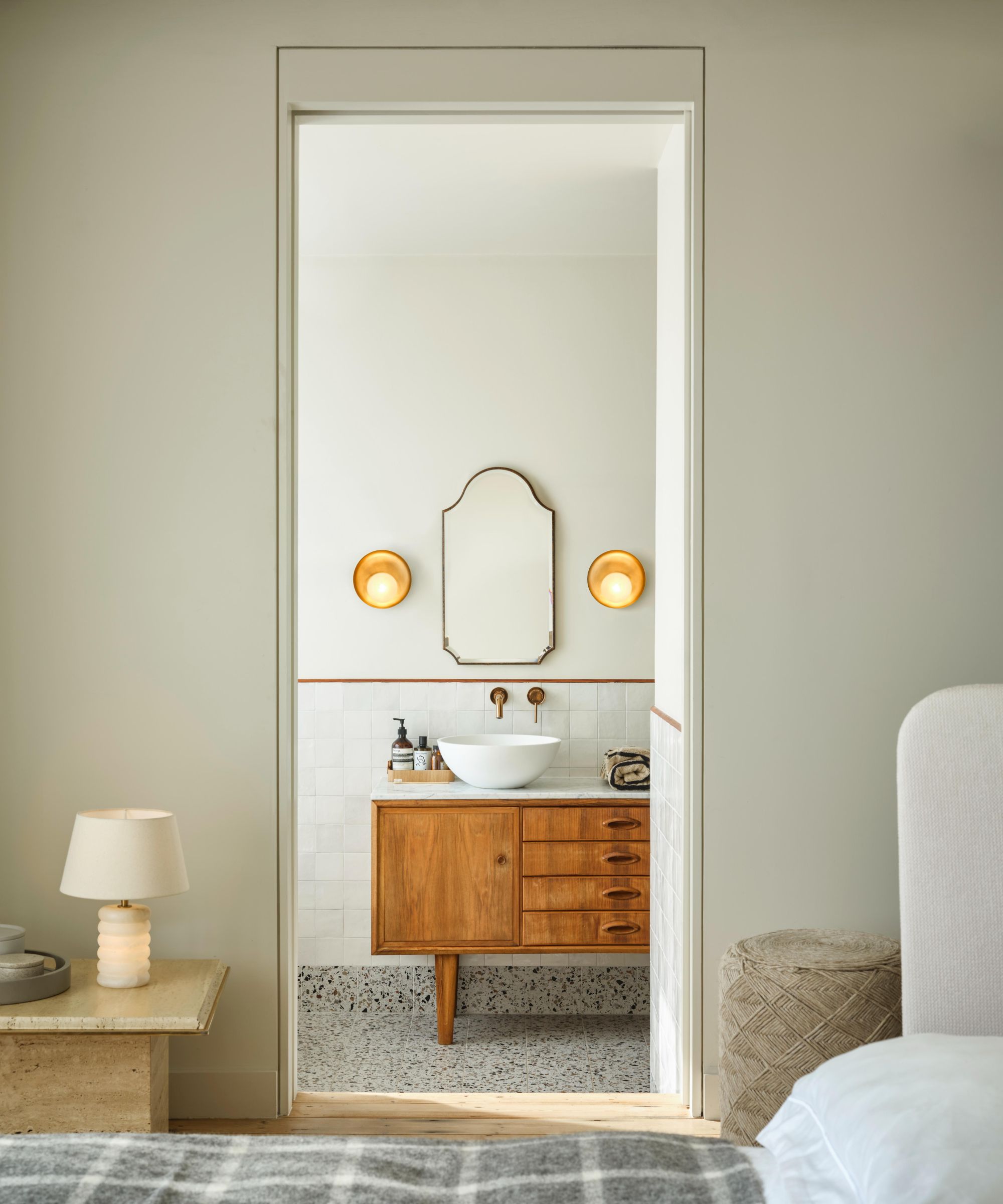A view through a bedroom doorway into a serene ensuite bathroom with white square tiles, a terrazzo floor and a mid-century style walnut vanity unit with a white vessel basin on a marble top. A brass wall-mounted tap, arched brass-framed mirror and two gold dome wall sconces complete the warm, elegant scheme.