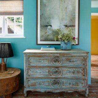 hallway wall decor ideas, bright blue hallway, with antique chest of drawers, terracotta floor tiles, artwork, pot with flowers, wooden side table and lamp