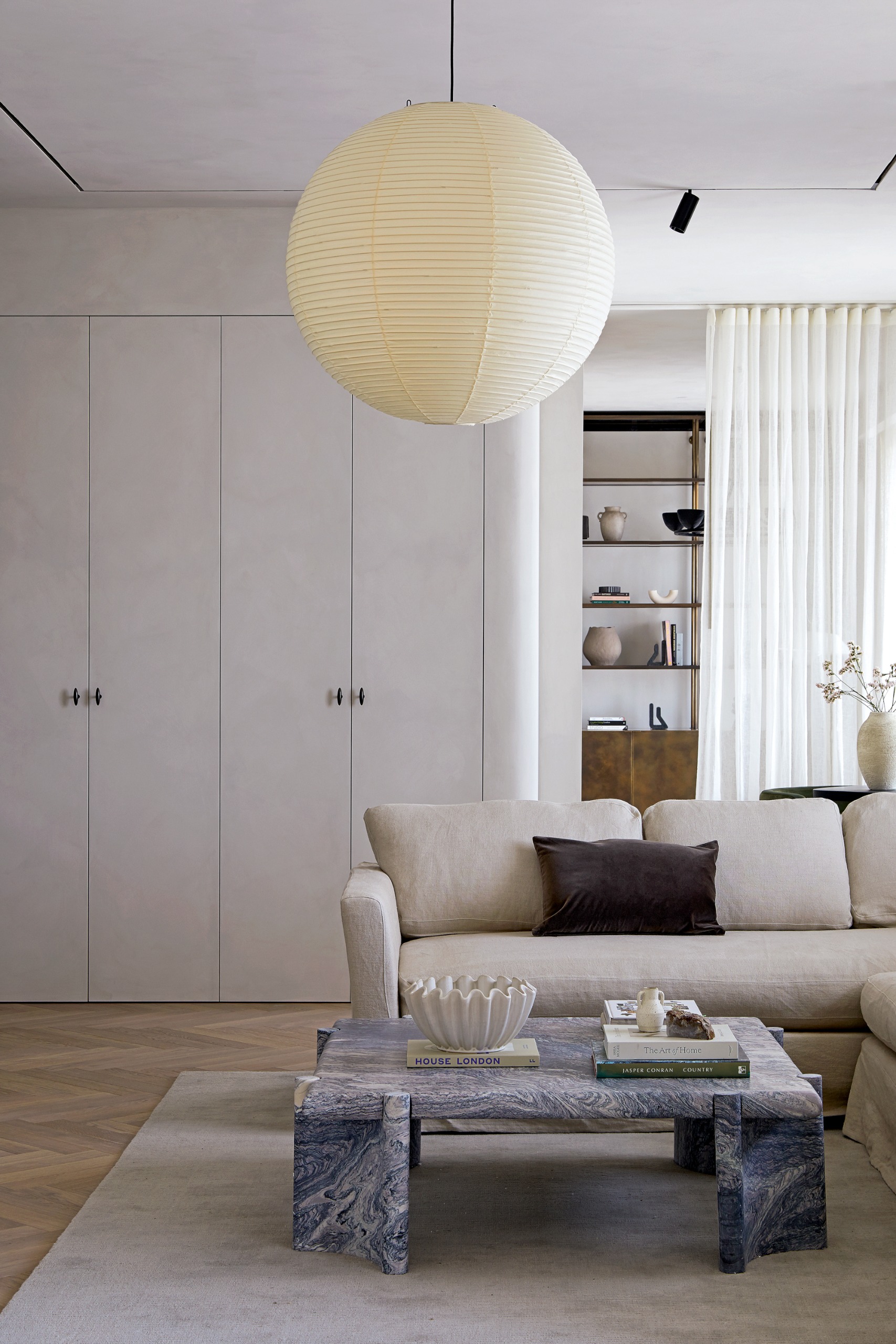 Living room with white walls and built-in cupboards, round paper pendant light, blue-gray marble coffee table, cream corner sofa and cream rug