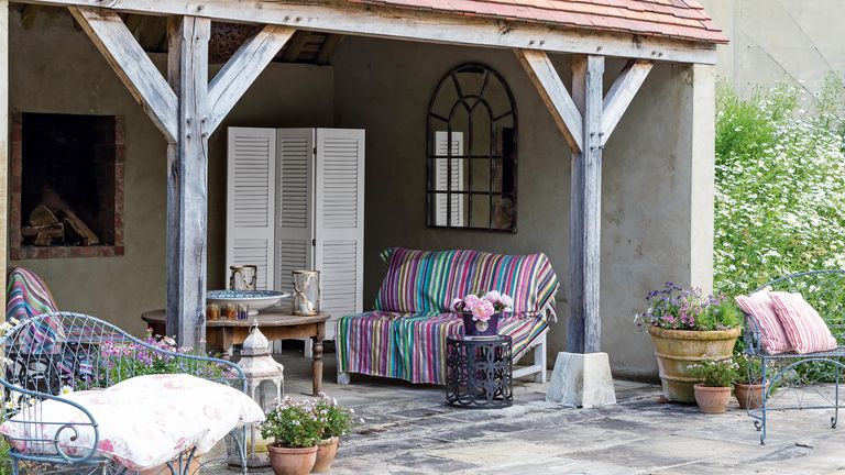 An example of outdoor living room ideas showing a terrace area with a roof, wooden beams and a sofa covered in a striped throw
