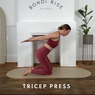 Lottie Anderson performs a triceps press. She wears matching leggings and sports bra and exercises on a Pilates mat in a home setting, in front of a fireplace and potted plant. She is kneeling, with her torso leaning forward at a 45 degree angle. Her arms are straight and held behind her.