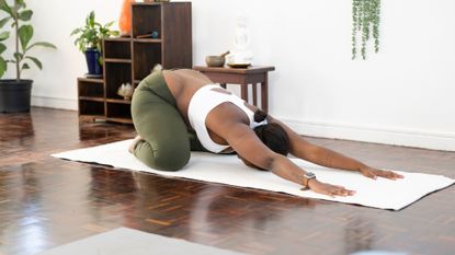 A woman wearing khaki green leggings and white crop top doing a childs pose in a living room