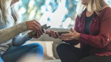 A woman counts out cash to another woman while they sit on the sofa together.