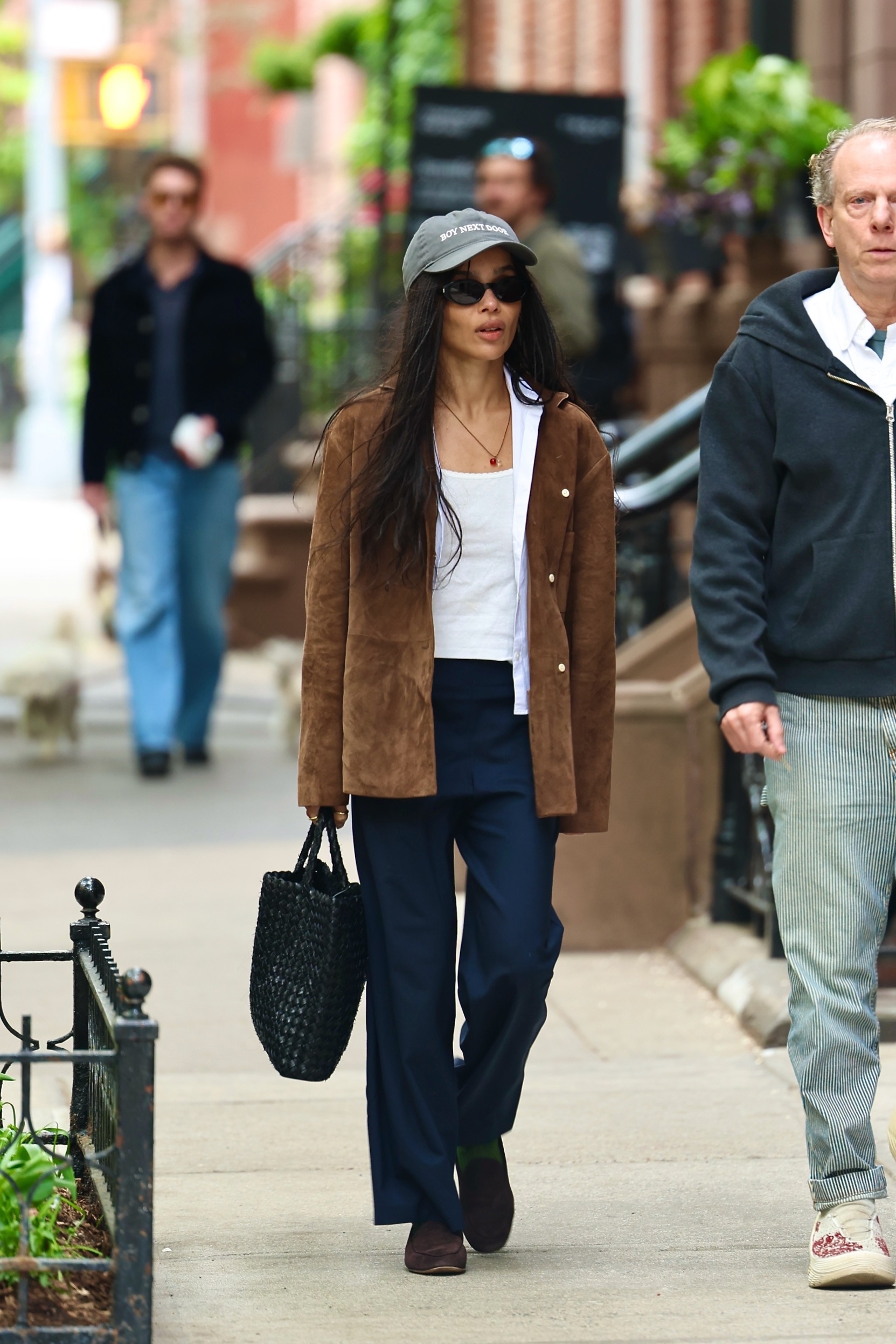 Zoe Kravitz wears a hat, suede jacket, white tee, trousers, loafers, and a raffia bag.