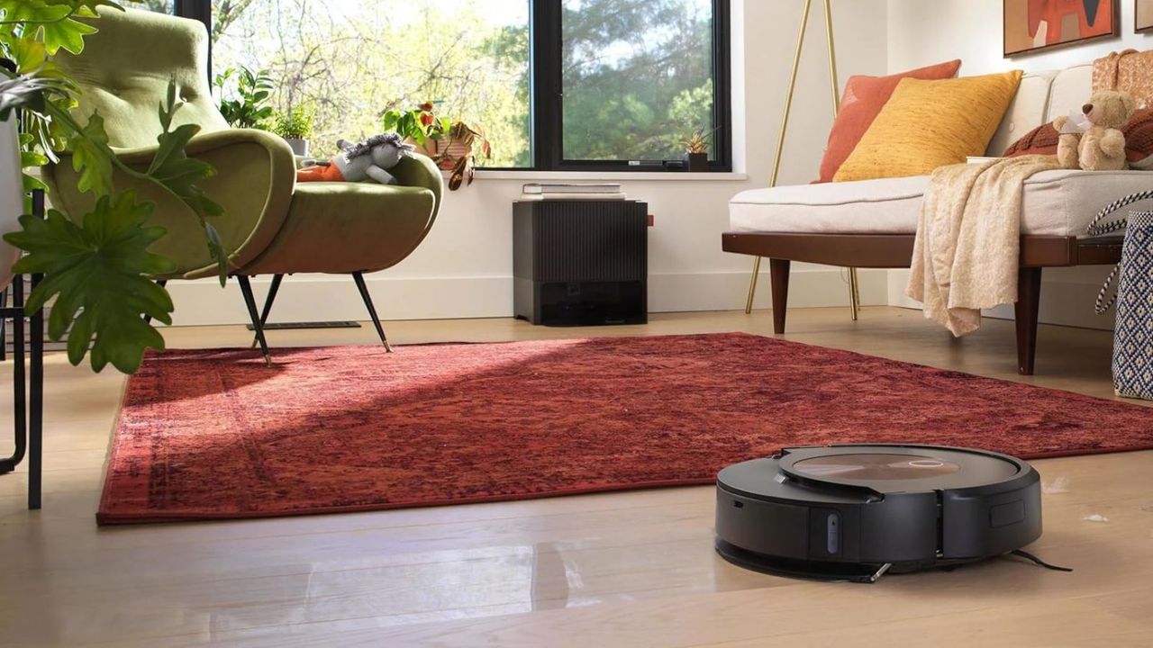 An iRobot Roomba j9+ Combo vacuuming a wooden floor in colorful living room with red rug, sofa, green armchair and plant