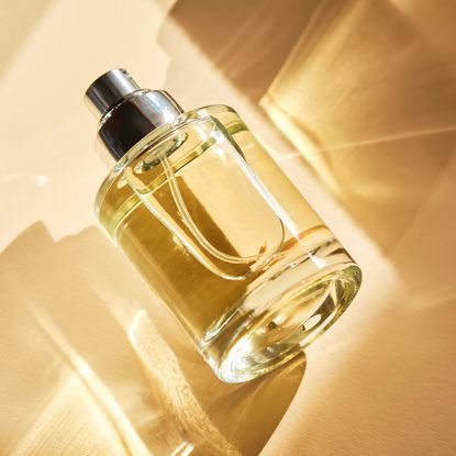 A perfume bottle with light reflecting through it against a cream background and surrounded by shadows