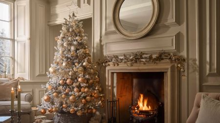 A creamy white living room with a tall flocked Christmas tree in minimalist white and peach baubles by a fireplace with a round mirror above