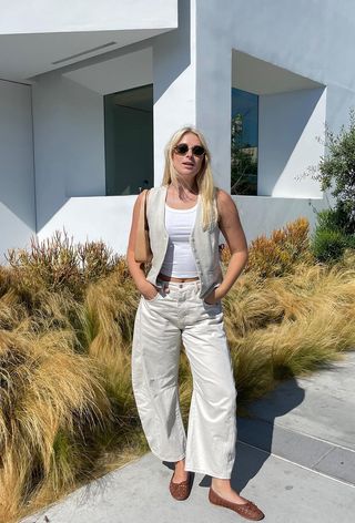 A woman standing outside wearing a tan waistcoat over a white tank top with white barrel-leg jeans and brown ballet flats.