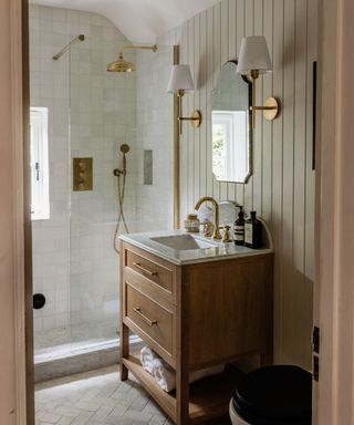 A small bathroom with a wooden standing sink with a marble counter, a mirror and two wall sconces above it, a shower with two gold shower heads, and a wooden door opening up to the room