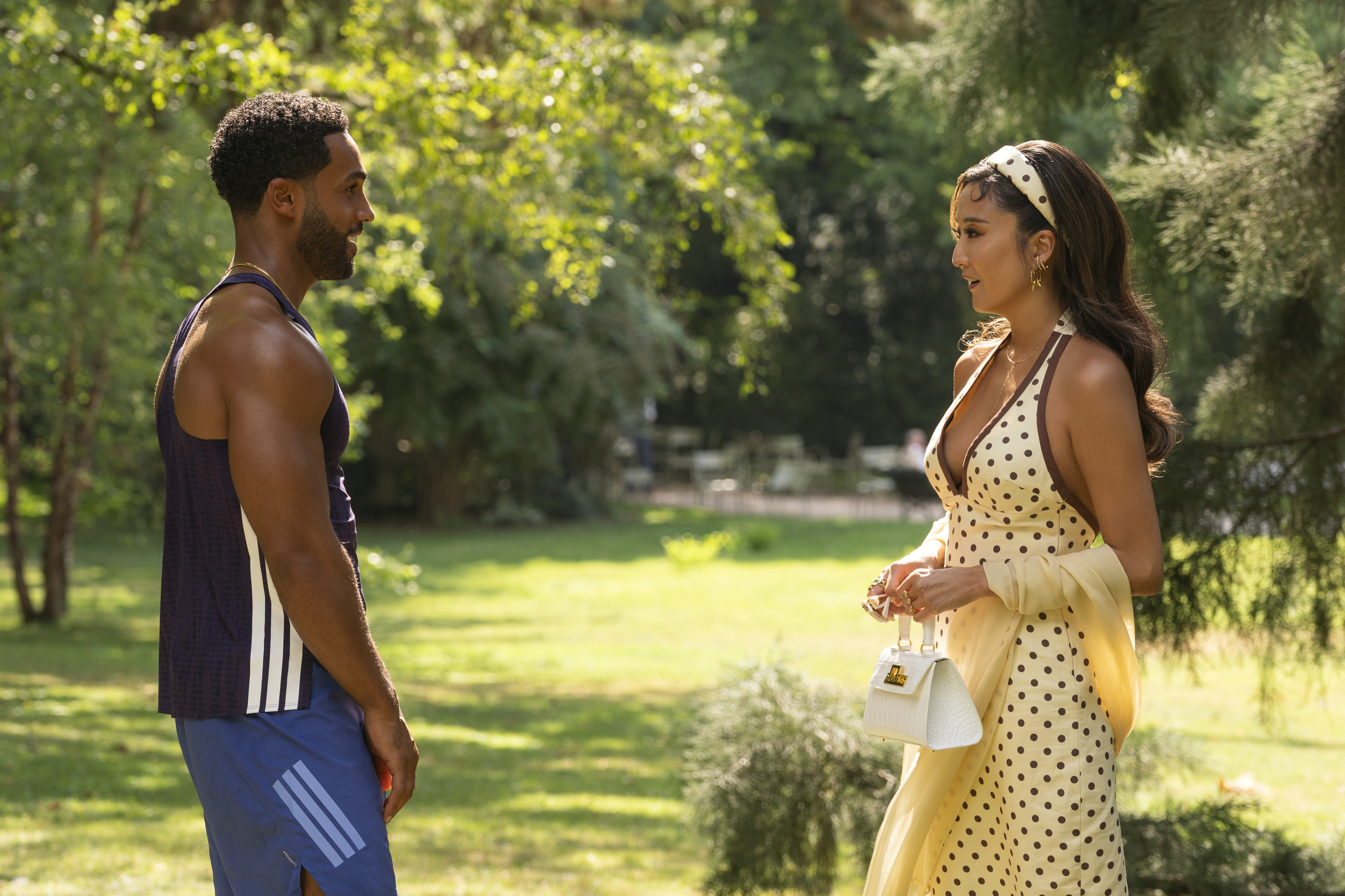 Lucien Laviscount as Alfie speaking to Ashley Park as Mindy in a sunny park in Emily in Paris season 6