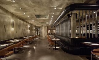 Alternative view of the dining area at Dash Kitchen featuring spotlights and stone coloured walls, ceiling and flooring. There are multiple dark brown tables and light brown chairs along with a curved row of black seats. The bar can be seen through the tall silver cylinders that separate the two areas