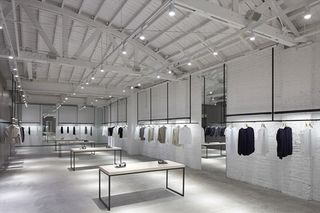 A fashion showroom featuring clothes items on hangers continuing around the perimeter of the room on rails; fashion accessories on four tables in the centre.