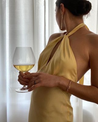 Fashion influencer Sasha Mei poses next to a window with white curtains wearing drop earrings from Lié Studio, a yellow satin halter dress, and gold chain bracelet while holding a glass of wine.