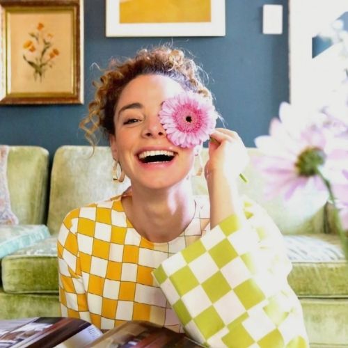 Image of a woman in a yellow and white checkerboard shirt sitting in front of a lime green sofa, posing with a pink flower in front of her face.