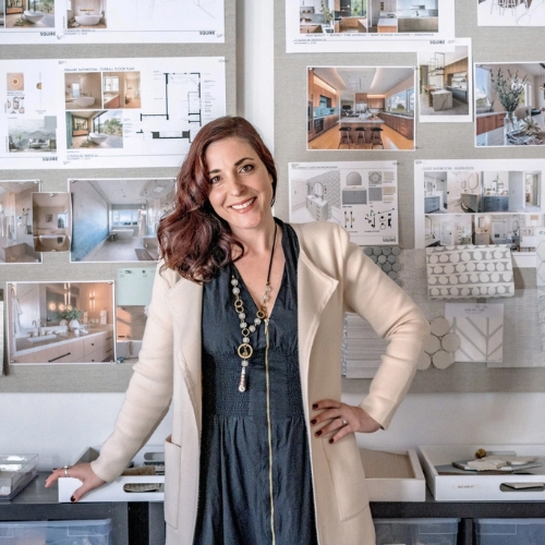 Image of a woman in a navy dress and cream jacket standing in front of a wall covered in design imagery.