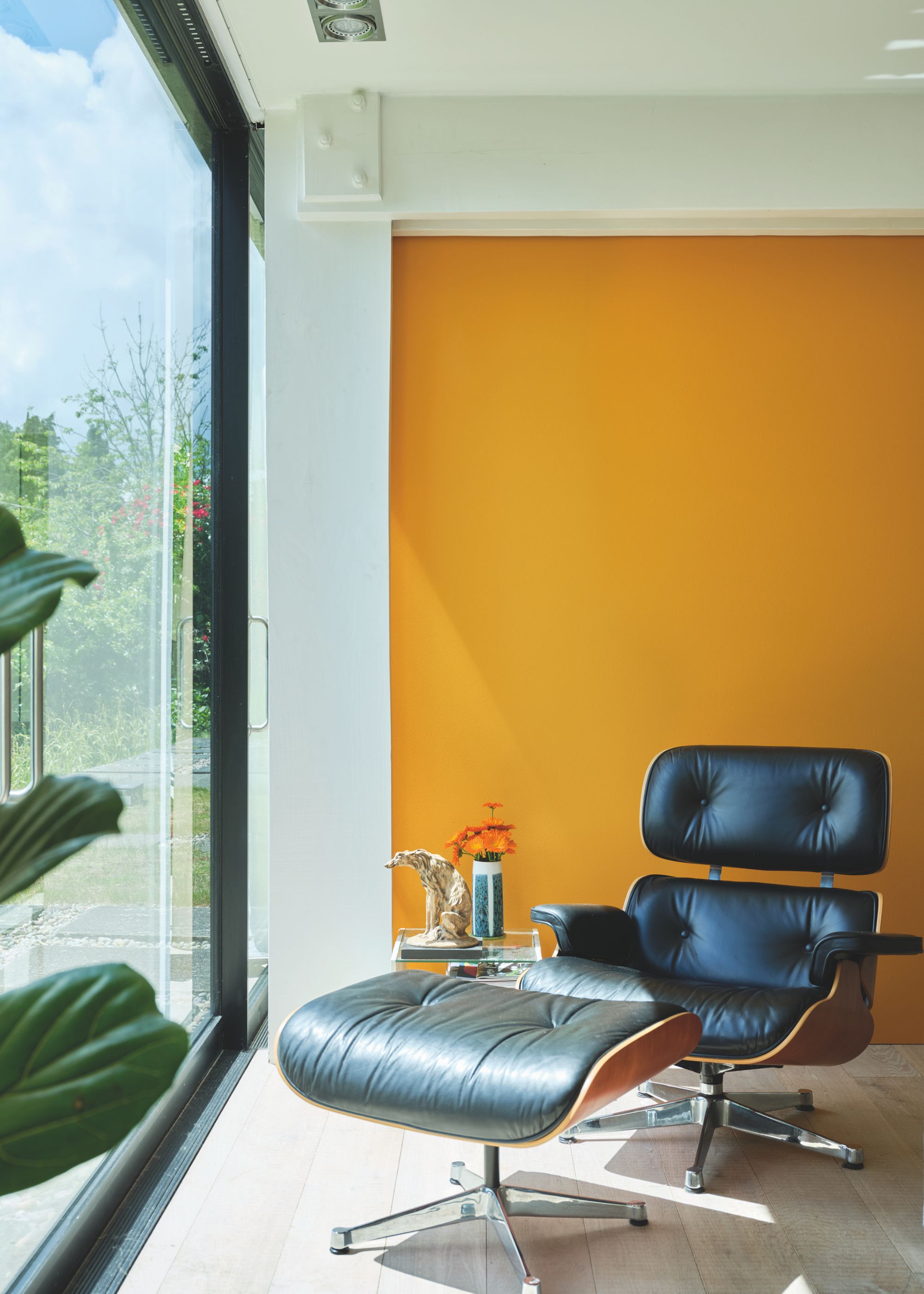 Bright corner of a living space with a large floor-to-ceiling window, a black leather chair with a footstool, and a bright orange accent wall.