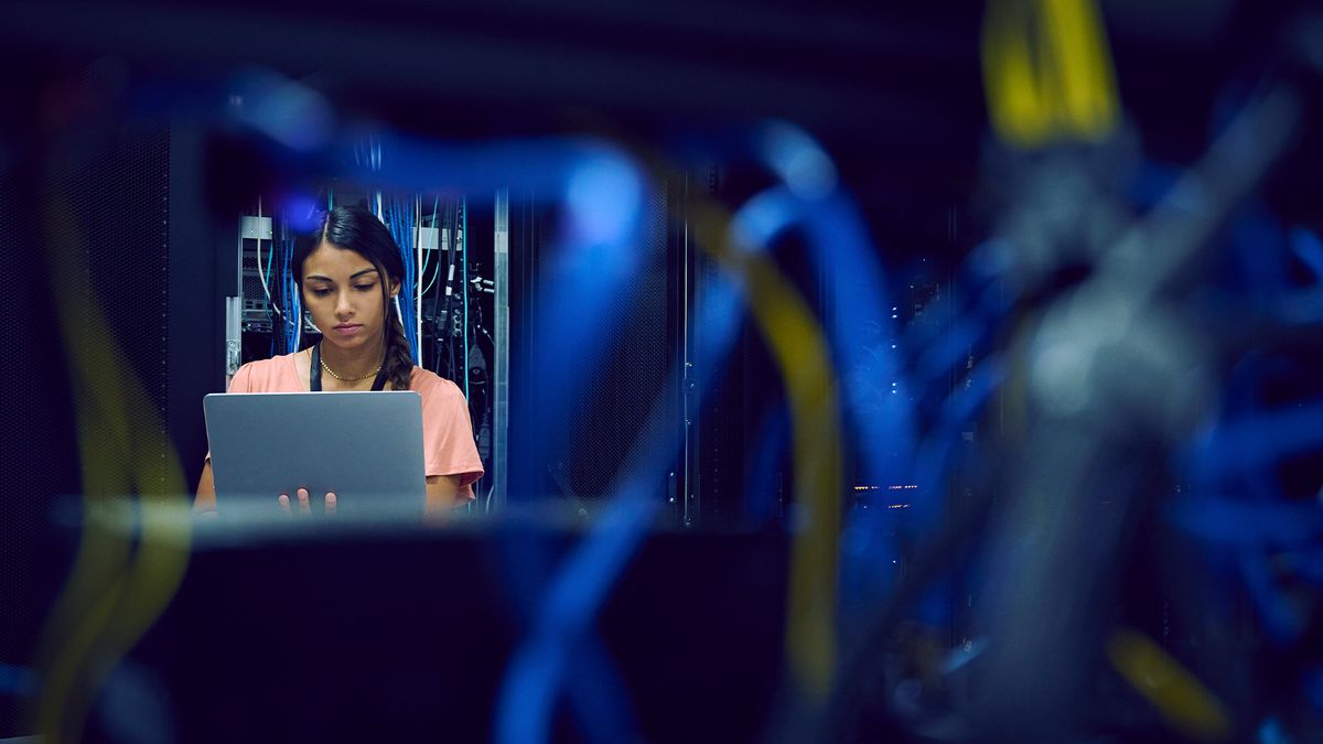 Cyber security worker at a desk in a server room with blurred wires and cabling in the foreground.