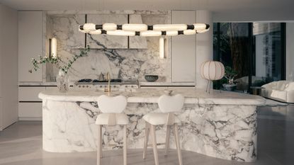 Image of an all white, ultra modern kitchen with a curved kitchen island made out of white marble. The backsplash and hood are in a matching marble and there is an abstract pendant light hanging over the island.