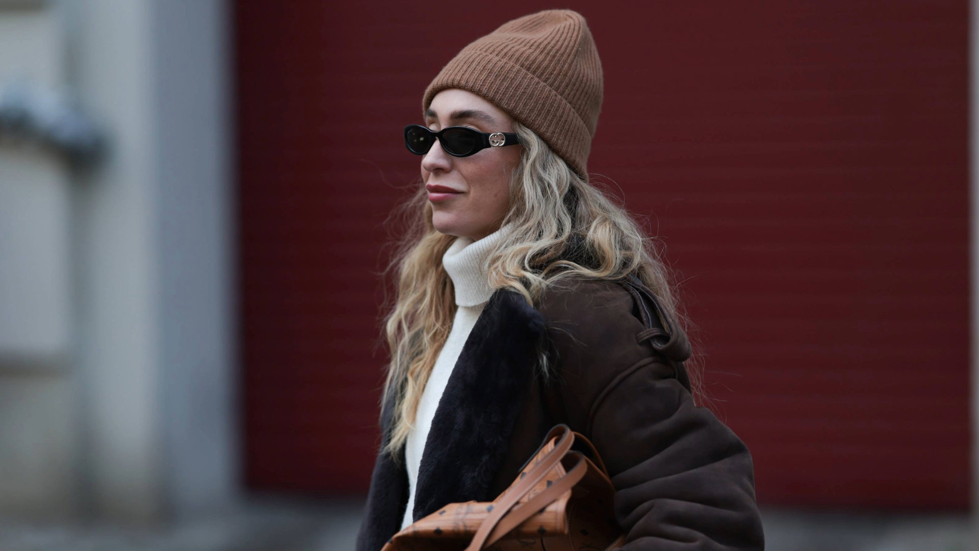 BERLIN, GERMANY - NOVEMBER 24: Sonia Lyson seen wearing a long dark brown shearling Ducie coat layered over wide, flowing cream trousers from Review and COS. The look is completed with soft slip-on shoes from Zara and a ribbed beige COS beanie. Accessories include a structured tan MCM tote bag with monogram patterning and slim black Gucci sunglasses. The outfit blends cozy winter textures with elevated neutral tones on November 24, 2025 in Berlin, Germany. (Photo by Jeremy Moeller/Getty Images)