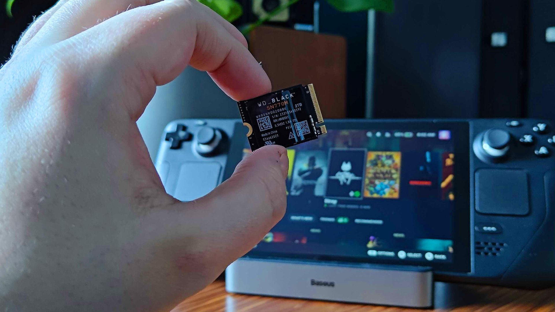 Hand holding WD Black SN770M SSD with Steam Deck in backdrop