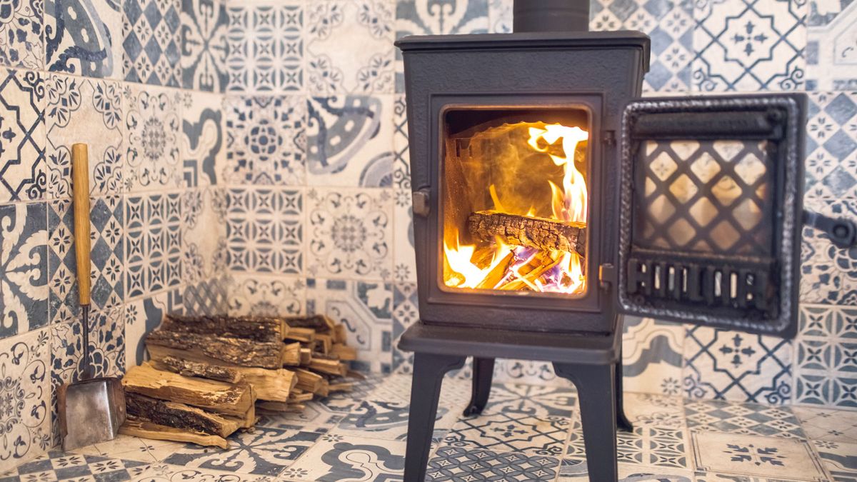 Lit black log burner against decorative tiled background with logs in corner