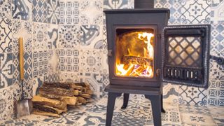 Lit black log burner against decorative tiled background with logs in corner