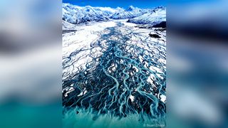 Aerial view of a glacial river meandering through a snowy landscape, surrounded by towering mountains and blue sky