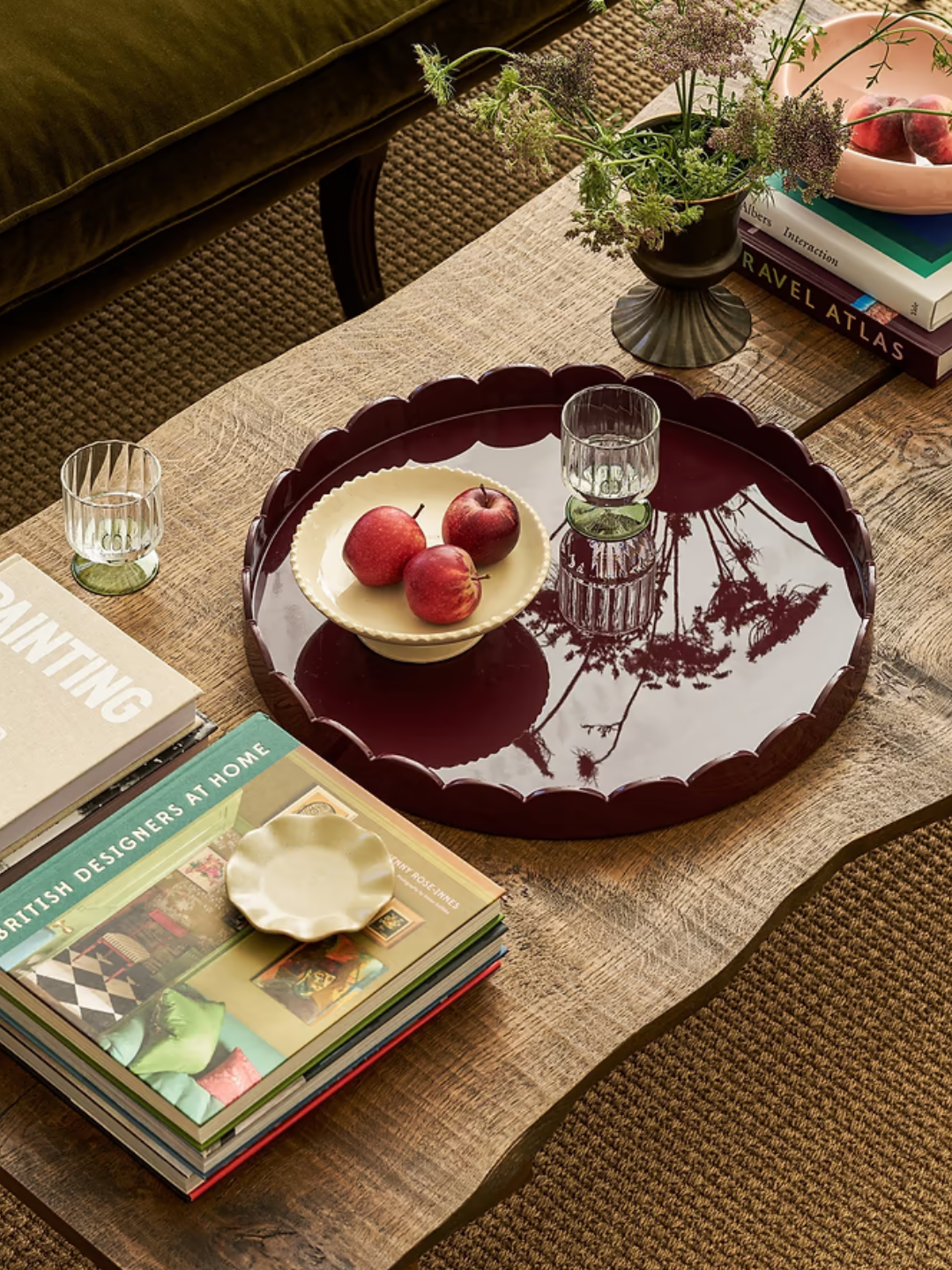 A scalloped maroon tray from Addison Ross on a living room table with a glass, a bowl of apples, a vase, and coffee table books