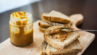 jar of peanut butter with a spoon, on a wooden board with slices of toast