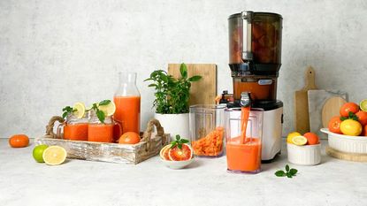 The Kuvings Auto 10 S - Hands Free Juicer on a marble counter against a matching backsplash. The juicer in action juicing citrus fruits, surrounded by various white bowls of oranges and lemons. A wooden tray with a carafe and two drinking mason jars filled with orange juice.