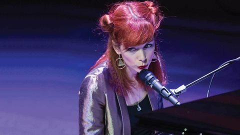 The Anchoress wearing a silver suit sitting behind a piano