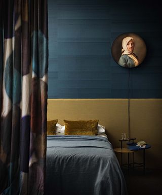 A dark blue bedroom with teal textured wallpaper, yellow ochre headboard and circular portrait painting on the wall.