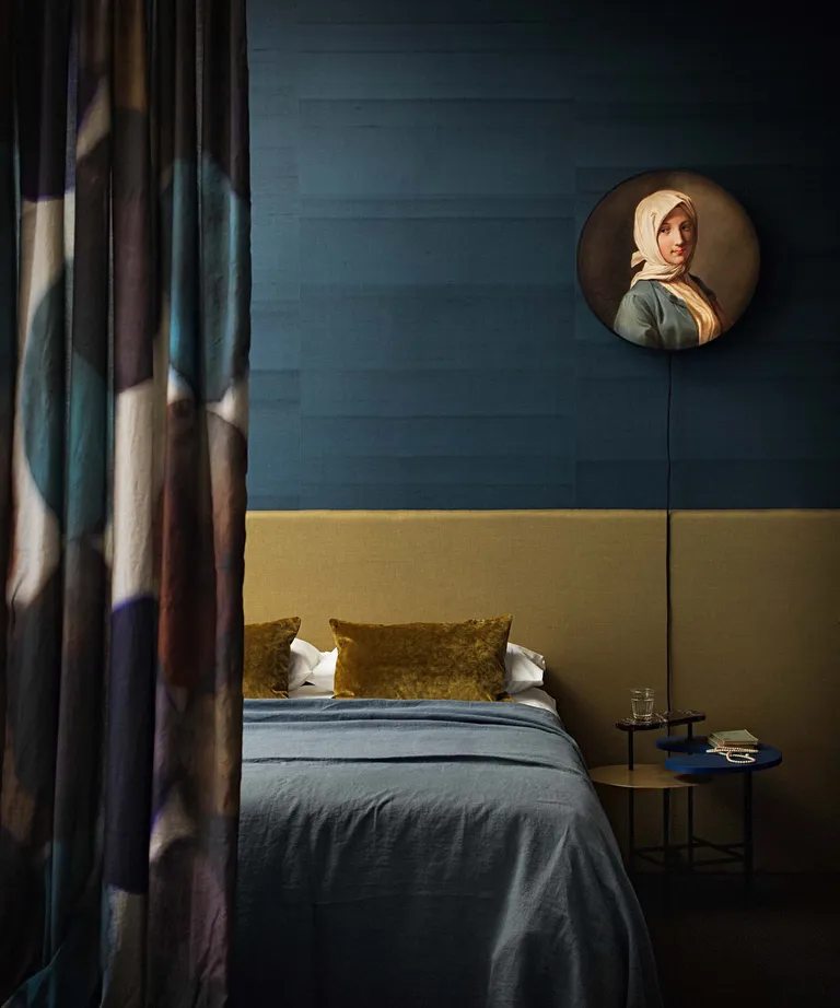 A bedroom with teal textured wallpaper, yellow ochre headboard and circular portrait painting on the wall