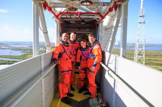 four people in orange spacesuits smile on a narrow walkway high above the ground