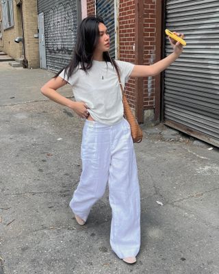 fashion influencer Mimi Nguyen waling in NYC wearing a beige T-shirt, white linen pants, a tan woven tote bag, and beige mesh flats