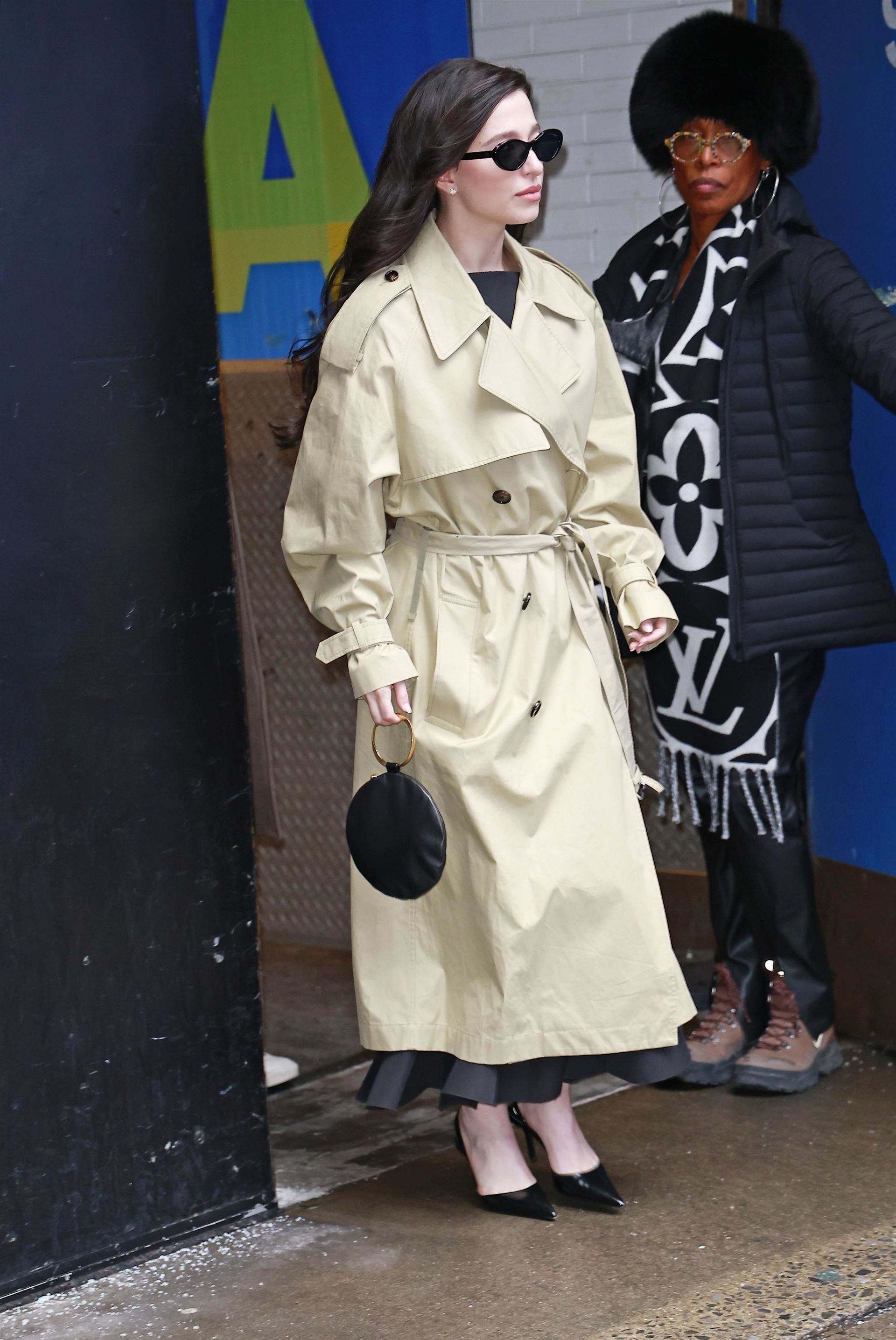 Mikey wears a trench coat, black pumps, black sunglasses, and a black ring-handle bag.