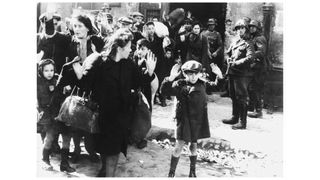 A black and white photograph taken in the Warsaw Ghetto in 1943 during World War II. It shows Jews, both adults and children, held at gunpoint as SS troops look on.
