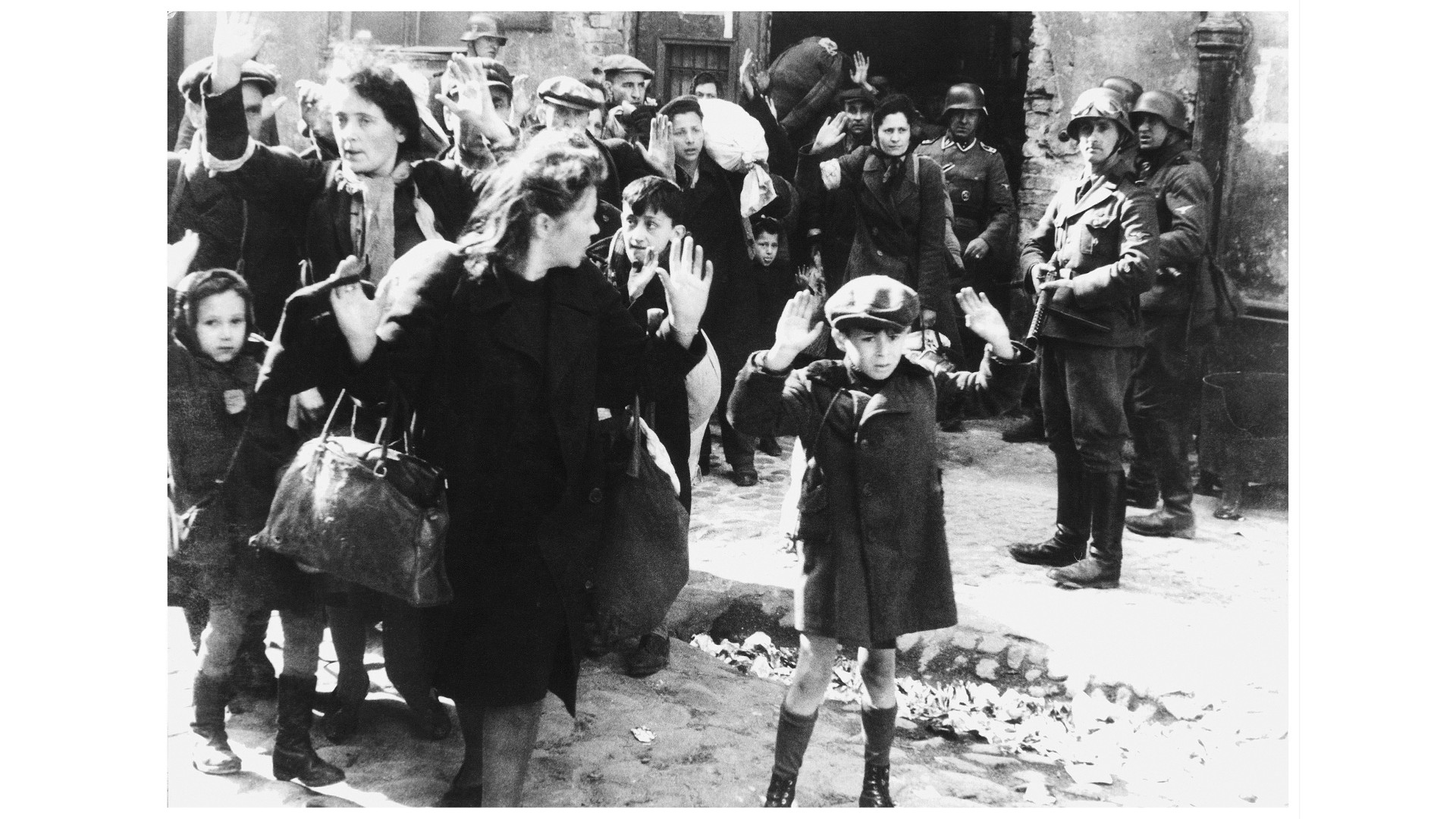 A black and white photograph taken in the Warsaw Ghetto in 1943 during World War II. It shows Jews, both adults and children, held at gunpoint as SS troops look on.