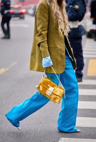 a close street-style photo of a woman wearing one of the most popular vintage bags, Fendi Baguette in yellow sequin styled with a green blazer, blue pants, and white shoes