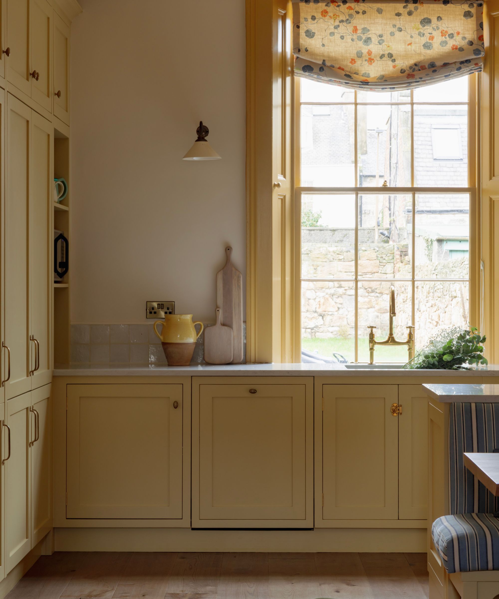 Georgian kitchen with yellow Shaker cabinets, brass hardware, grey stone countertops, and a sash window with floral Roman blind.
