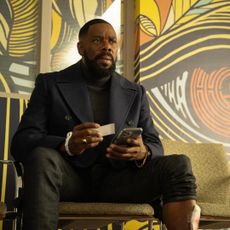 Colman Domingo as Muncie Daniels, holding a business card and a phone while sitting in front of a wall with a mural of an eye-shaped maze, in Episode 101 of The Madness.